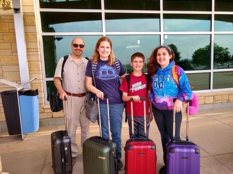 An adult man and woman stand with a pre-teen boy and girl. All of them have a backpack on their backs and a small suitcase in front of them.