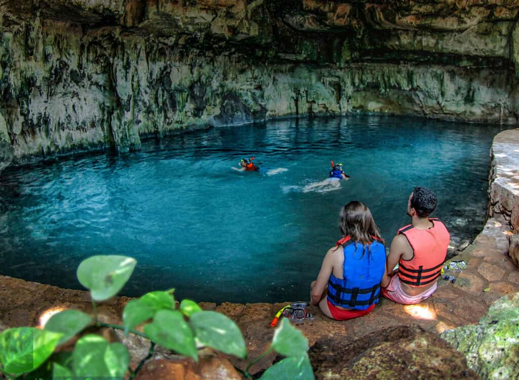 Two people sit on the edge of the Carlotta cenote at Hacienda Mucuyche