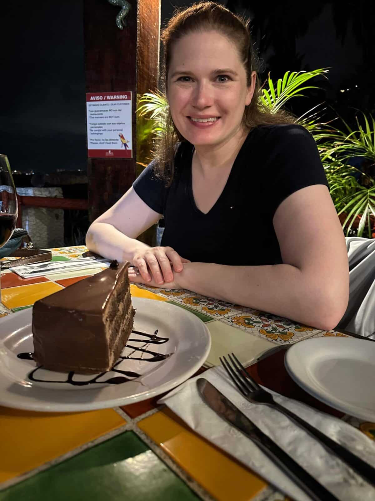 A woman sits at a table with a piece of chocolate cake in front of her.