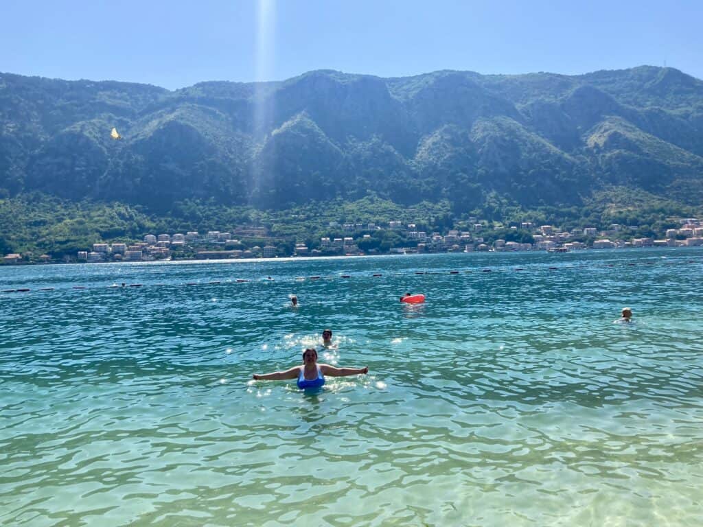 Two teens swimming in blue water with green mountains behind them at Kotor Beach